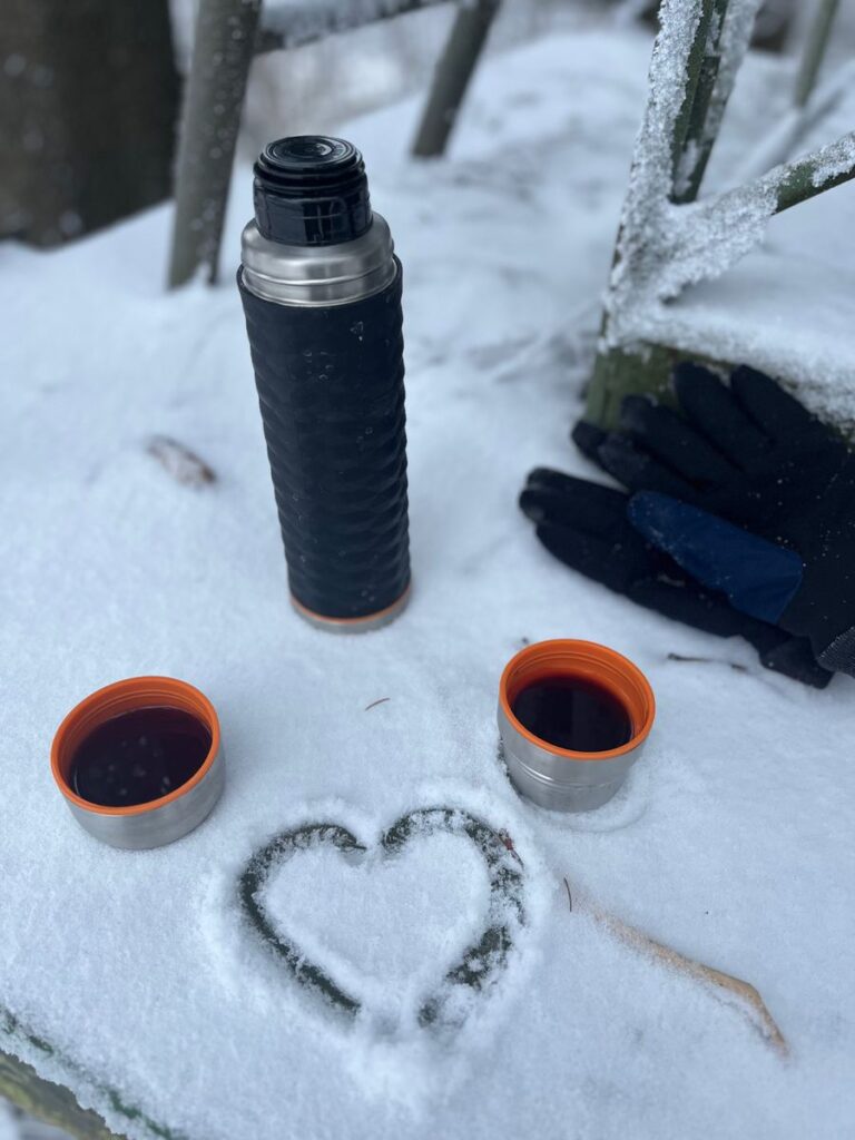 Thermoskanne und zwei Becher Glühwein im Schnee, davor ein in den Schnee gemaltes Herz