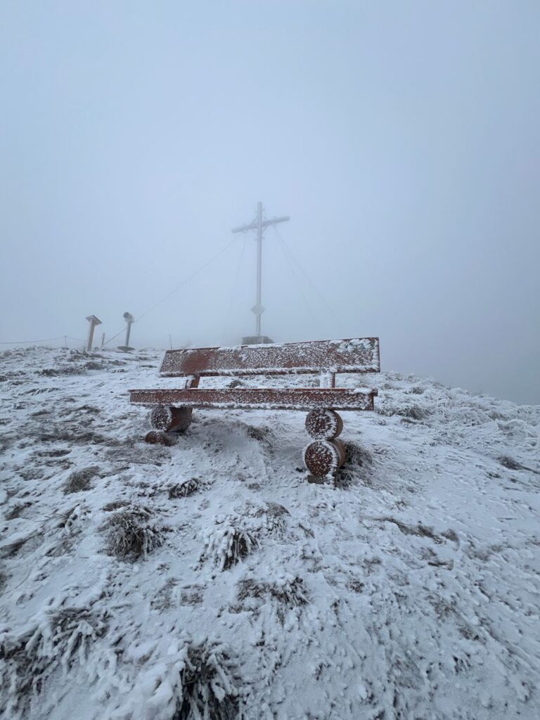 Vereistes Gipfelkreuz und Bank mit Raureif auf dem Pferdskopf im dichten Winternebel