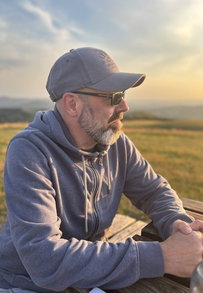 Steffen von FuldaRhön sitzt im Abendlicht an einem Holztisch auf der Wasserkuppe, im Hintergrund die Rhön