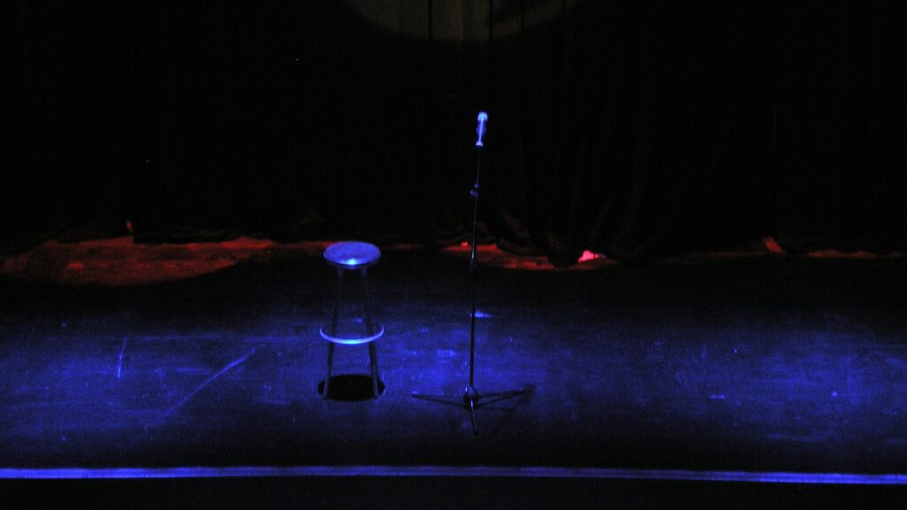 Leere Comedy-Bühne im blauen Licht: ein Barhocker und ein Mikrofonständer vor schwarzem Vorhang