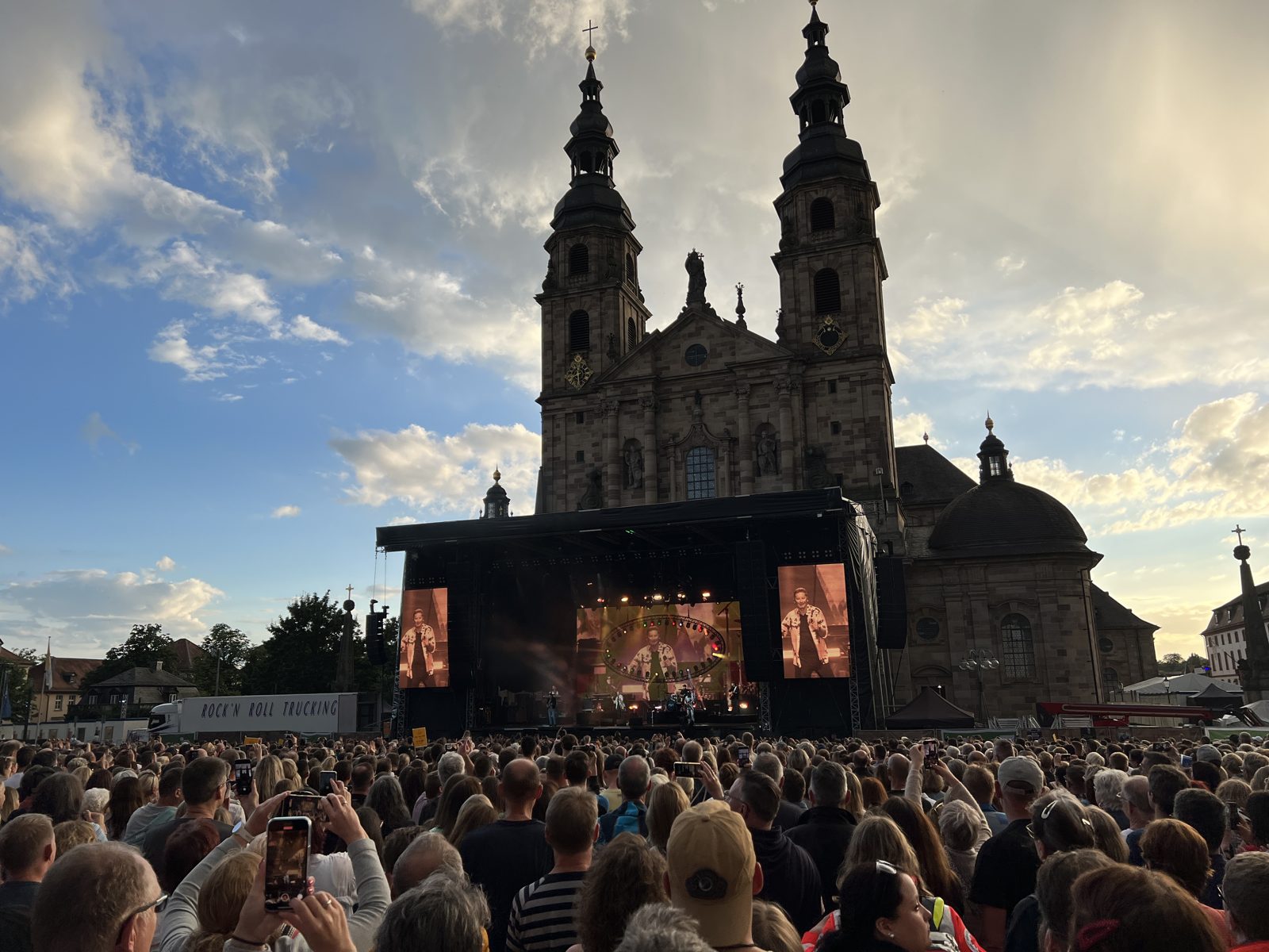 Open-Air-Bühne vor dem Fuldaer Dom bei einem Domplatzkonzert, davor das Publikum, darüber Abendhimmel mit Wolken.