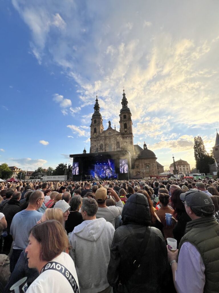 Besucher eines Domplatzkonzerts in Fulda, dahinter Bühne und Dom unter einem Abendhimmel.
