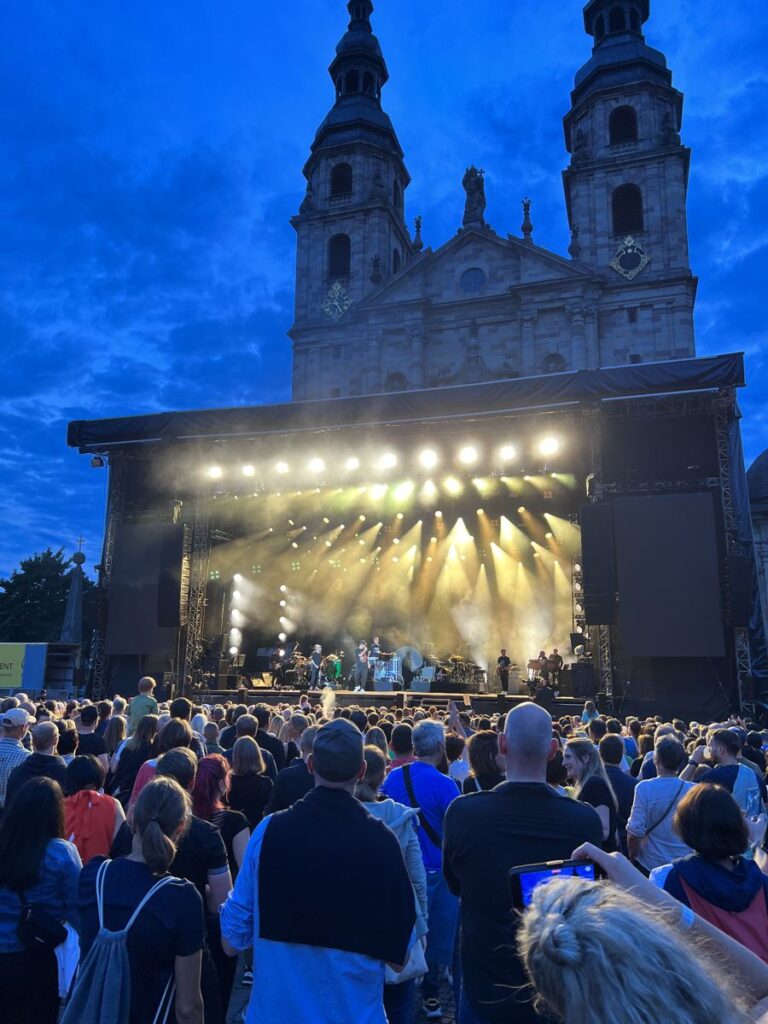 Publikum vor der beleuchteten Bühne eines Domplatzkonzerts in Fulda, im Hintergrund der Dom bei Dämmerung.