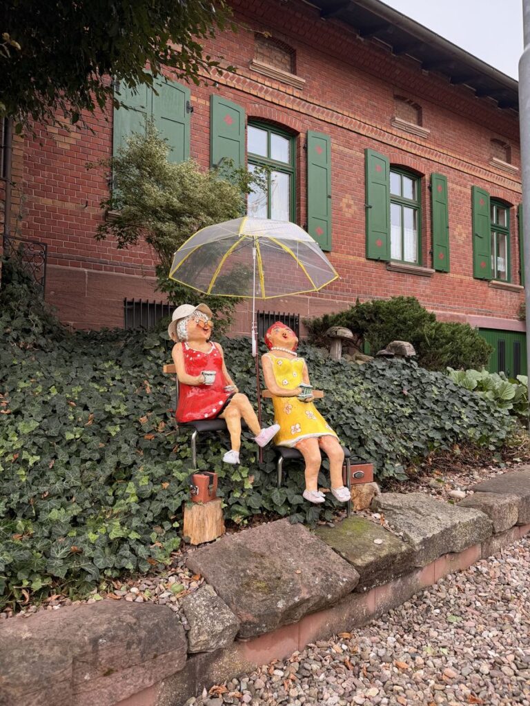 Zwei bunte Holzfiguren unter einem Regenschirm im Vorgarten eines Backsteinhauses in Geisa