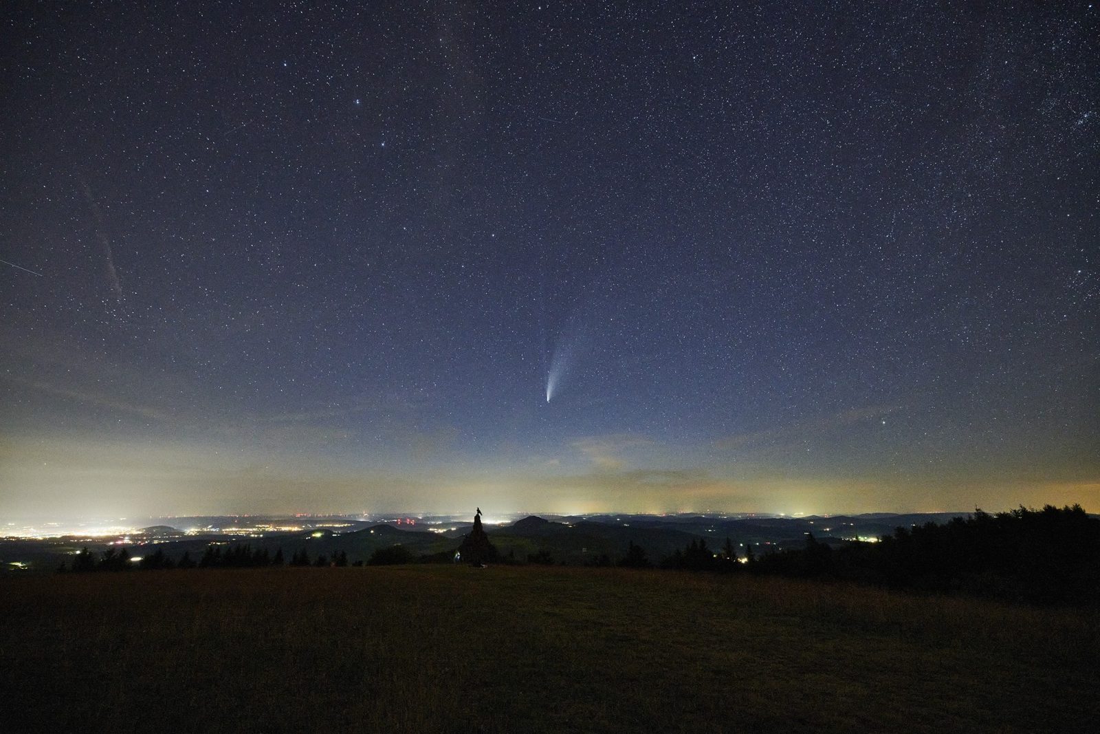 Ein Komet mit Schweif am Nachthimmel, darunter der helle Lichtschein von Orten am Horizont.