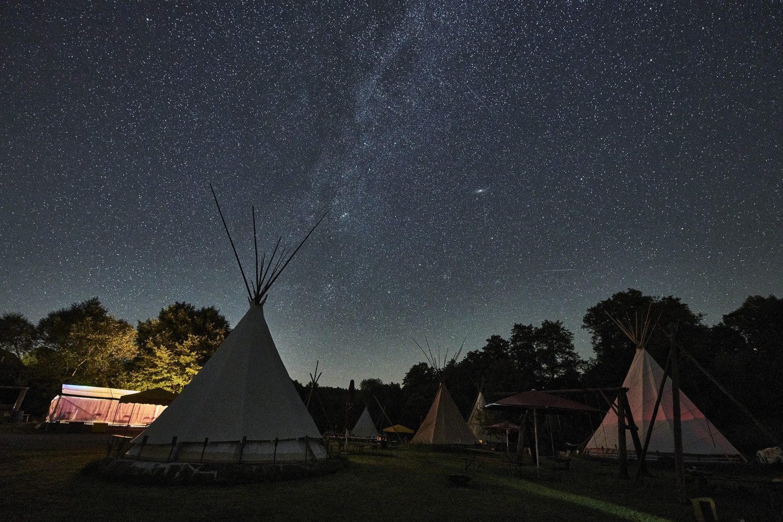 Die Milchstraße steht über mehreren Tipi-Zelten, am Rand ein paar Bäume.