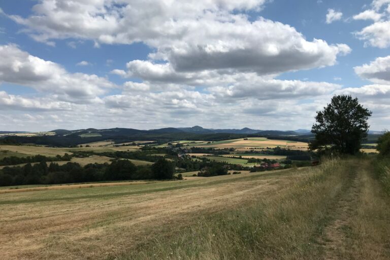 Weiter Blick über Felder und Wiesen auf die Kuppen der Rhön unter Sommerwolken