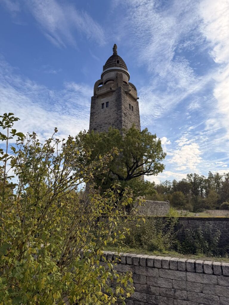 Wittelsbacher Turm hinter Bäumen und einer Natursteinmauer vor blauem Himmel